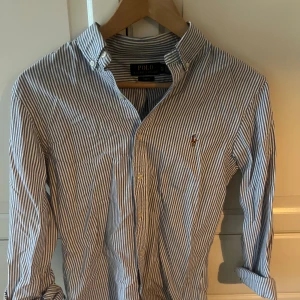 Randig skjorta från Polo Ralph Lauren - Snygg slim fit skjorta från Polo Ralph Lauren i blått och vitt med klassiskt randigt mönster. Skjortan har knappar framtill, button-down krage och det ikoniska broderade logot på bröstet. Perfekt för dig som gillar stilrena och tidlösa plagg. Pris kan diskuteras.
