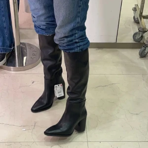 Svarta höga stövlar med klack - Säljer ett par svarta höga stövlar i skinn med snygg spetsig tå och klack. Stövlarna går upp till knäet och har en stilren, minimalistisk design som passar till jeans eller kjol. Använda ett par gånger men fortfarande som nyskick. Ca 8cm klack. 100% skinn, från Zara, nypris: 1800kr. 