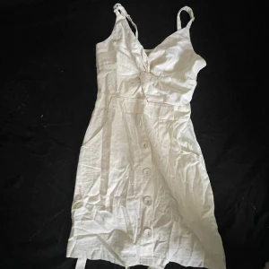 Vit kort klänning från Jasmine - Supersöt vit kort klänning från Jasmine med smala axelband, knyt i midjan och knappar framtill. Klänningen har v-ringad omlott-topp och är gjord i ett lätt linneliknande material, perfekt för varma dagar.