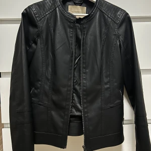 Svart jacka från Soyaconcept - Snygg svart jacka i konstläder från Soyaconcept med raka linjer och quiltade detaljer på axlarna. Jackan har dragkedja framtill och två fickor med dragkedja. 