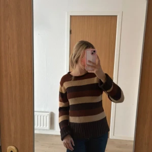 Randig stickad tröja i brunt och beige - Säljer en stickad tröja med breda ränder i mörkbrunt, mellanbrunt och beige. Tröjan har rund hals, långa ärmar och ribbade muddar. Perfekt för dig som gillar retrovibbar och jordnära färger.