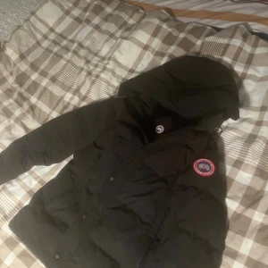 Svart dunjacka från Canada Goose - Svart dunjacka från Canada Goose med huva och klassisk logga på ärmen. Jackan är quiltad och har dragkedja samt tryckknappar framtill. Perfekt för kalla dagar och har en clean, stilren look som funkar till det mesta.