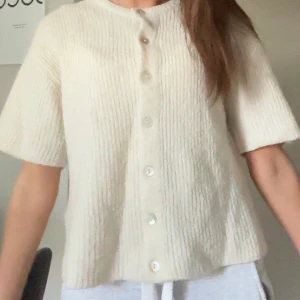 Krämvit kofta från Gina Tricot - Superfin krämvit kofta från Gina Tricot i stickat material. Koftan har korta ärmar, rund halsringning och knäppning med pärlemorknappar framtill. Stl s, lite nopprig med fof i bra skick 