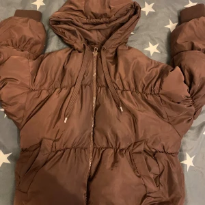 Brun pufferjacka från H&M Divided XS - Säljer en brun pufferjacka från H&M Divided i storlek XS. Jackan har huva med snörning, dragkedja framtill och två fickor. Oversized känsla med puffiga ärmar och ribbade muddar. Perfekt för kalla dagar när du vill vara både varm och snygg.