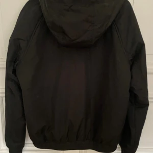 Svart vindjacka från Calvin Klein Jeans - Svart vindjacka från Calvin Klein Jeans med huva, dragkedja framtill och två stora fickor. Jackan har logga på bröstet och ärmen, samt elastisk nederkant. Perfekt för dig som vill ha en clean och stilren look.