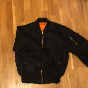 Svart bomberjacka från Alpha Industries - Säljer en svart bomberjacka från Alpha Industries med klassisk orange insida. Jackan har dragkedja framtill, fickor med tryckknappar och en ärmficka med dragkedja. Ställ gärna frågor om intresserad!