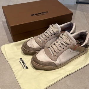 Burberry sneakers i vitt och beige - Snygga sneakers från Burberry i vitt och beige skinn med klassiskt rutigt mönster på sidorna. Skorna har rund tå, platt sula och snörning framtill. Perfekta för dig som gillar en stilren och lyxig look med en touch av brittisk design. Nypris 7599kr mitt pris 1899kr