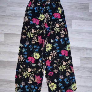 Blommiga vida byxor från H&M - Snygga svarta byxor från H&M i storlek 36 med färgglatt blommönster i blått, rosa, gult och beige. Vida ben och skön passform med resår och snörning i midjan. Perfekta för dig som gillar en avslappnad och trendig stil.