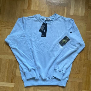 Ljusblå Stone Island sweatshirt - Säljer en ljusblå sweatshirt från Stone Island med klassisk rund hals och ribbade muddar. Tröjan har den ikoniska Stone Island-loggan på ärmen och är tillverkad i mjuk bomull. Perfekt för dig som gillar stilrena och trendiga plagg.