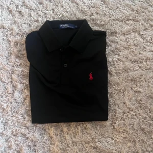 Svart pikétröja från Ralph Lauren - Klassisk svart pikétröja från Ralph Lauren med röd broderad logga på bröstet. Tröjan har krage, knäppning med två knappar och är kortärmad. Perfekt för en clean och stilren look.