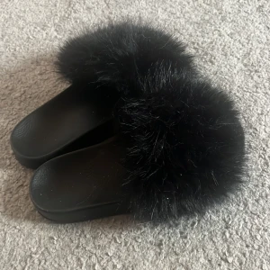 Svarta sandaler med fuskpäls - Svarta sandaler med bred rem täckt av mjuk och fluffig fuskpäls. Snygg och chill look som passar perfekt till sommaren eller när du vill vara extra cozy hemma. Platt sula och enkel slip-on-modell gör dem superenkla att ta på.