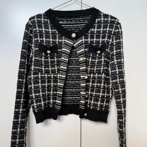 Svartvit rutig kofta med pärlknappar - Snygg svartvit rutig kofta med långa ärmar och dekorativa pärlknappar framtill. Koftan är stickad och har två fejkfickor på bröstet. Perfekt för dig som vill ha en trendig och chic look med lite extra detaljer.