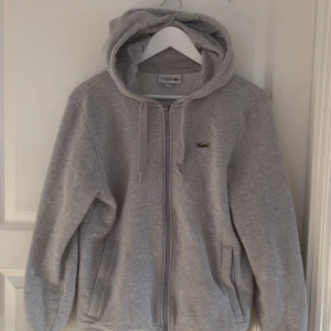 Grå hoodie från Lacoste Sport - Säljer en grå hoodie från Lacoste Sport med dragkedja och huva. Tröjan har snörning i huvan, två fickor framtill och den klassiska gröna krokodilloggan på bröstet. Perfekt för chill dagar eller när du vill ha en sportig look.