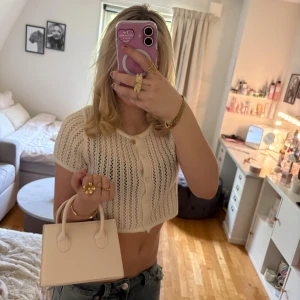 Vit minihandväska i skinnimitation - Supersöt liten handväska i vit skinnimitation med stilren, fyrkantig form och två handtag. Perfekt storlek för det viktigaste och har guldfärgade detaljer på sidorna för axelrem. Enkel och clean design som passar till många outfits. Och remmen medkommer såklart.