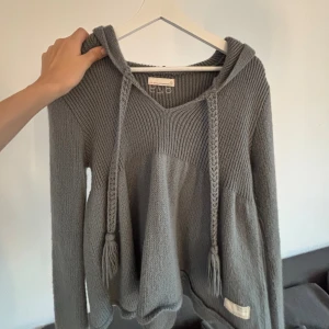 Odd Molly grå stickad hoodie med snörning - Mysig grå stickad hoodie från Odd Molly med långa ärmar, v-ringning och flätade snören med tofsar. Tröjan har en avslappnad passform och huva, perfekt för lager på lager under höst och vinter. Snygg detalj med liten etikett nertill.