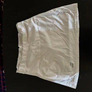 Vit kortkjol från Babolat - Säljer en vit sportig kortkjol från Babolat, perfekt för tennis. Kjolen har en enkel och stilren design med elastisk midja. Materialet är lätt och syntetiskt, vilket gör den skön att röra sig i på banan.