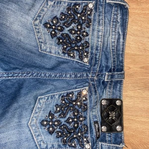 Blå Miss Me jeans med broderade fickor - Snygga blå jeans från Miss Me med broderade bakfickor och glittriga detaljer. Modellen har låg midja, raka ben med slits framtill och dekorativa nitar. Jeansen är i stretchigt denim och har coola slitningar för en trendig look.