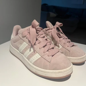 Rosa campus - Snygga Adidas sneakers i ljusrosa mocka med klassiska vita ränder på sidorna och vit sula. Skorna har rund tå, breda matchande skosnören och Adidas logga bak på hälen. Perfekta för dig som vill ha en stilren och trendig look.