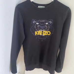 Svart Kenzo sweatshirt med tigertryck - Svart sweatshirt från Kenzo med ikoniskt broderat tigerhuvud och gult Kenzo Paris-tryck på bröstet. Tröjan har rund hals, långa ärmar och ribbade muddar. Perfekt för dig som vill sticka ut med en streetstyle-look. Passar S/xs tun tröja 