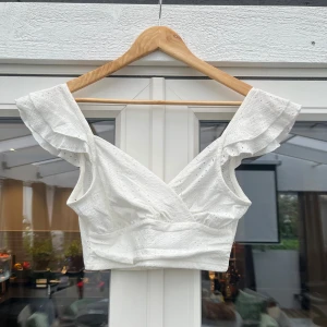 Vit omlottopp med volangärmar - Superfin vit omlottopp med broderade detaljer och volangärmar. Toppen är croppad och har en v-ringad skärning som ger en snygg siluett. Perfekt för varma dagar och somriga outfits.