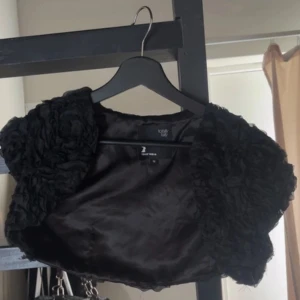 Svart bolero - Kort svart boleroväst från Tally Weijl med glansig finish och dekorativa blommiga volanger över hela ytan. Västen har öppen front och är perfekt för att ge outfiten en extra touch. Snygg och unik design som sticker ut.