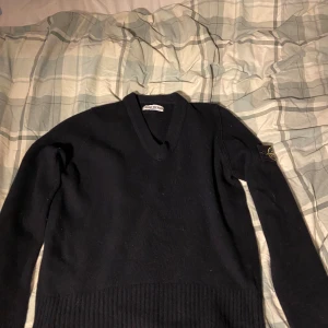 Svart v-ringad tröja från Stone Island - Svart v-ringad tröja från Stone Island med klassisk patch på ärmen. Tröjan är ribbstickad nedtill och vid ärmsluten, tillverkad i 80% ull och 20% nylon. Perfekt för dig som gillar stilrena och ikoniska plagg.