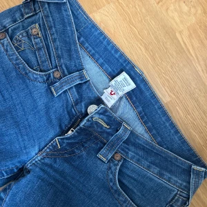Blå True Religion jeans bootcut - Säljer ett par klassiska blå jeans från True Religion med snygga kontrastsömmar och ikonisk broderad bakficka. Modellen har låg midja och bootcut-ben, tillverkade i mjukt bomullsjeansmaterial. Perfekta för dig som gillar en avslappnad men ändå trendig look.