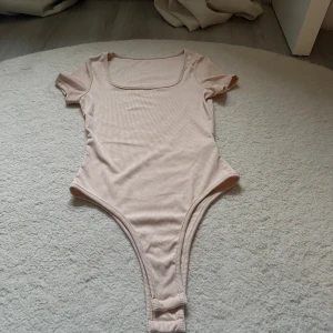 Ljusbeige ribbad bodysuit med kort ärm - Supersnygg ljusbeige bodysuit med ribbad struktur och korta ärmar. Den har en fyrkantig urringning och är tillverkad i mjukt, stretchigt material som sitter tight. Perfekt att styla med jeans eller kjol för en clean look.
