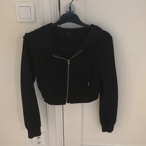 Svart croppad hoodie med dragkedja - Svart croppad hoodie med dragkedja framtill och huva. Tröjan har långa ärmar, ribbade muddar och snörning i huvan. Perfekt för en chill och stilren look.