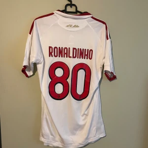 AC Milan Ronaldinho 80 Adidas tröja - AC Milan bortatröja med Ronaldinho 80 på ryggen. Vit med röda och svarta detaljer, broderat klubbmärke och guldstjärna på bröstet. Adidas-logga och Bwin sponsortryck. Tillverkad i lätt och ventilerande polyester (Clima365).