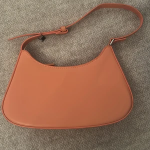 Orange axelväska i syntet - Trendig orange axelväska med stilren, minimalistisk design och justerbart axelband. Väskan har en mjukt rundad form och är tillverkad i slätt syntetmaterial. Perfekt för dig som vill addera en färgklick till din outfit.