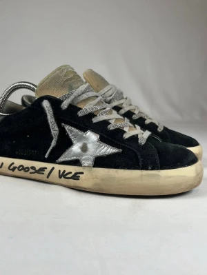 Golden Goose sneakers med silverstjärna - Svarta Golden Goose sneakers med ikonisk silverfärgad stjärna på sidan och handskriven text på den slitna sulan. Skorna har snörning, rund tå och är tillverkade i mocka och skinn. Tungan är beige och har en vintage-look. Perfekta för dig som gillar streetstyle.
