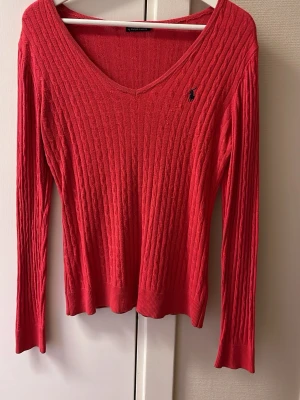 Röd stickad tröja från Ralph Lauren - Säljer en snygg röd stickad tröja från Ralph Lauren med v-ringad hals och kabelstickat mönster. Tröjan har långa ärmar och ett litet broderat logotyp på bröstet. Perfekt för dig som gillar klassisk stil med en modern touch.