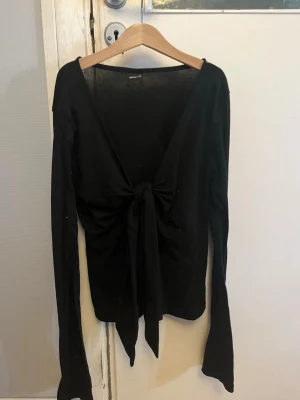 Gina top - Svart långärmad topp med djup v-ringning och snygg knytning framtill. Tunn och lätt i materialet, perfekt för att styla med jeans eller kjol. Enkel men trendig design som ger en cool vibe.