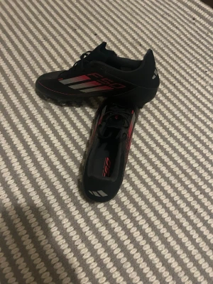 Adidas F50  fotbollsskor svarta - Säljer ett par Adidas F50 fotbollsskor i storlek 46. Skorna är svarta med röda och silvriga detaljer, har snörning och metall-dobbar för extra grepp på planen. Snygg och sportig design med klassiska Adidas-ränder på sidan. Perfekt för dig som vill ha grymma skor till matchen.