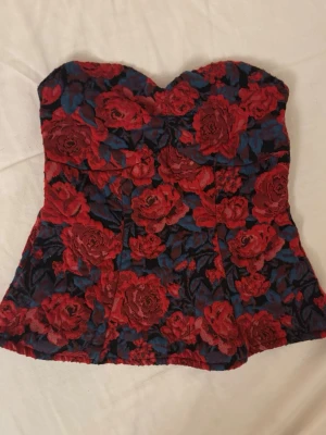 Blommig Topp från Gina Tricot XS - Säljer en snygg Top från Gina Tricot i storlek XS (mindre i storleken). Toppen är axelbandslös och har ett blommigt mönster med röda rosor mot en svart och mörkblå bakgrund. Perfekt för dig som vill sticka ut med färg och mönster.