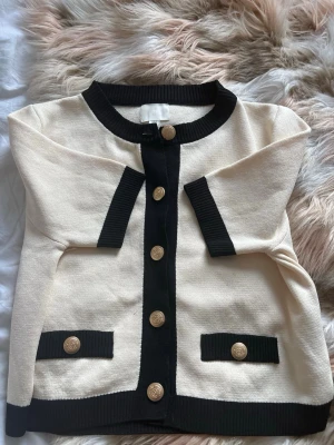 Beige och svart kofta från H&M - Snygg beige kofta från H&M med svarta ribbade detaljer och guldknappar framtill. Koftan har långärmade ärmar och två dekorativa fickor med knappar. Materialet är mjukt och stickat, fick av en kompis som aldrig fick någon användning av den o har själv aldrig använt den.💘