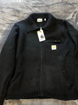 Svart stickad kofta från Carhartt - Svart stickad kofta från Carhartt med klassisk krage och dragkedja framtill. Har en liten ficka med dragkedja på bröstet och Carhartt-logga i gult och vitt. Ribbstickade muddar vid ärmslut och nederkant. Perfekt för lager-på-lager under kyliga dagar.