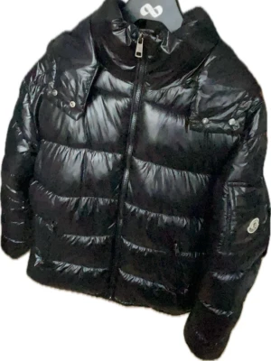 Svart pufferjacka från Moncler - Svart moncler jacka med glansig yta och klassisk logga på ärmen. Jackan har dragkedja framtill, fickor med lock och knappdetaljer. Perfekt för kalla dagar och har en riktigt snygg, modern look.