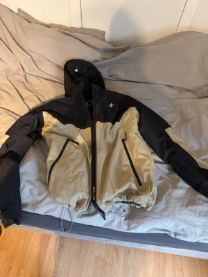 Beige och svart skidjacka Cross Sportswear XL - Snygg skidjacka från Cross Sportswear i beige och svart med huva och dragkedja framtill. Jackan har två stora fickor med dragkedja, justerbar nederkant med snörning. Perfekt för dagar i backen och outdoor-aktiviteter.
