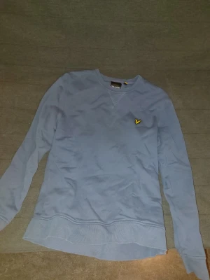 Blå långärmad tröja från Lyle & Scott - Snygg blå långärmad tröja från Lyle & Scott med klassisk rund hals och gul broderad logga på bröstet. Tröjan är i mjuk bomull och har ribbade muddar vid ärmslut och nederkant. Perfekt för chill dagar och enkel att matcha med jeans.