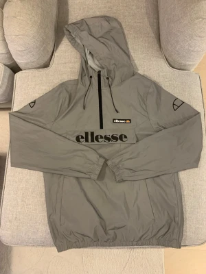 Grå vindjacka från Ellesse med huva - Snygg grå vindjacka från Ellesse i storlek L men känns som M. Jackan har huva med snörning, half zip framtill och elastiska muddar vid ärmar och nederkant. Stor Ellesse-logga på bröstet och mindre loggor på ärmarna. Perfekt för dig som gillar sportig stil.