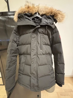 Carson Parka Canada Goose M - Säljer en svart dunjacka från Canada Goose med fluffig päls på huvan och klassisk logga på ärmen. Jackan har dragkedja och knappar framtill, flera fickor och är quiltad för extra värme. Perfekt för kalla vinterdagar och riktigt snygg stil.