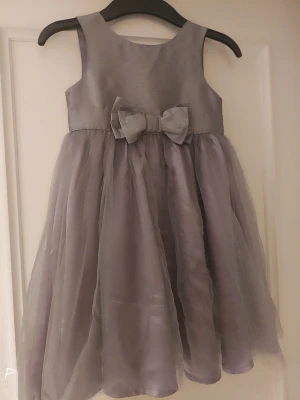 Grå ärmlös klänning med rosett ,4-5år - Supersöt grå klänning med voluminös kjol i tyll och en stor rosett framtill i midjan. Klänningen är ärmlös och har en rund halsringning. Perfekt för dig som gillar en elegant och stilren look med extra fluffig kjol.