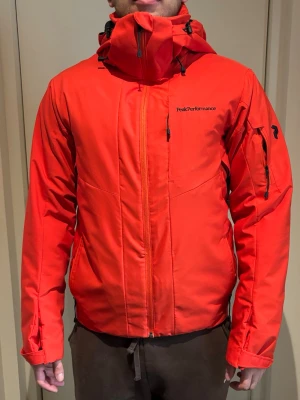 Röd Peak Performance skidjacka - Peak Performance skidjacka i stark röd färg med huva och flera dragkedjor, inklusive ventilationsdragkedjor under armarna och ficka på ärmen och snölås
