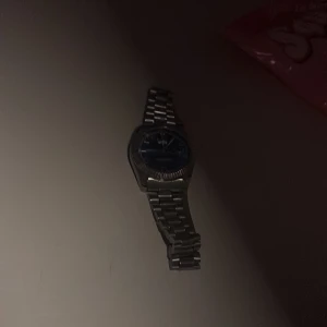 Silverfärgad klocka med blå urtavla - Stilren klocka med metallarmband i silver och rund blå urtavla. Klockan har en klassisk länkdesign och markerad boett. Perfekt för dig som gillar en clean och modern look. Urtavlan har tydliga visare och detaljer som ger en snygg kontrast mot det silvriga armbandet. Med en liten spricka vid glaset 