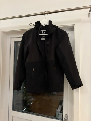 Svart skaljacka från Lindberg - Svart skaljacka från Lindberg med huva och dragkedja framtill. Jackan har två sidofickor med dragkedja och en diskret svensk flagga på bröstet. Märkespatch på ärmen och stilren design som funkar till många outfits.