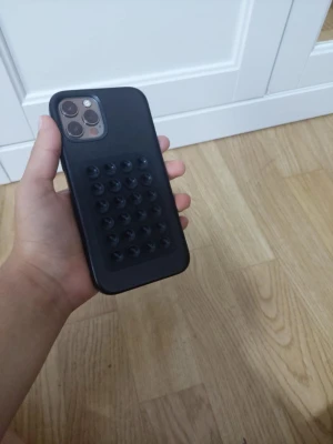 iPhone 12 Pro - Snygg iPhone 12 Pro med trippelkamera och svart skyddsfodral med sugproppar för extra grepp. Telefonen ser ut att vara i gott skick utan synliga repor eller skador. Perfekt för dig som vill ha en kraftfull och stilren mobil med extra skydd.