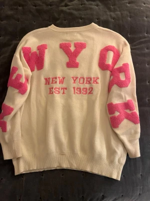 Beige stickad tröja LISSIMO med rosa detaljer - Säljer en beige stickad tröja från LISSIMO med stora rosa bokstäver och texten 'NEW YORK EST 1992' på ryggen. Tröjan har långärmade och loose passform, lite utanvänddd
