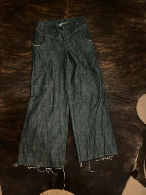 Blå wide jeans med rå kant - Säljer ett par blå jeans med breda ben och rå, fransig kant längst ner. Byxorna har klassiska fickor fram och ett coolt, lite slitet uttryck. Perfekt för dig som gillar avslappnad och trendig stil.